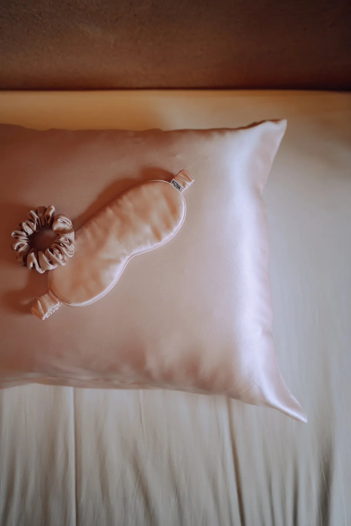A blush silk Kova pillowcase on a bed with a matching silk sleep mask and scrunchie resting on top