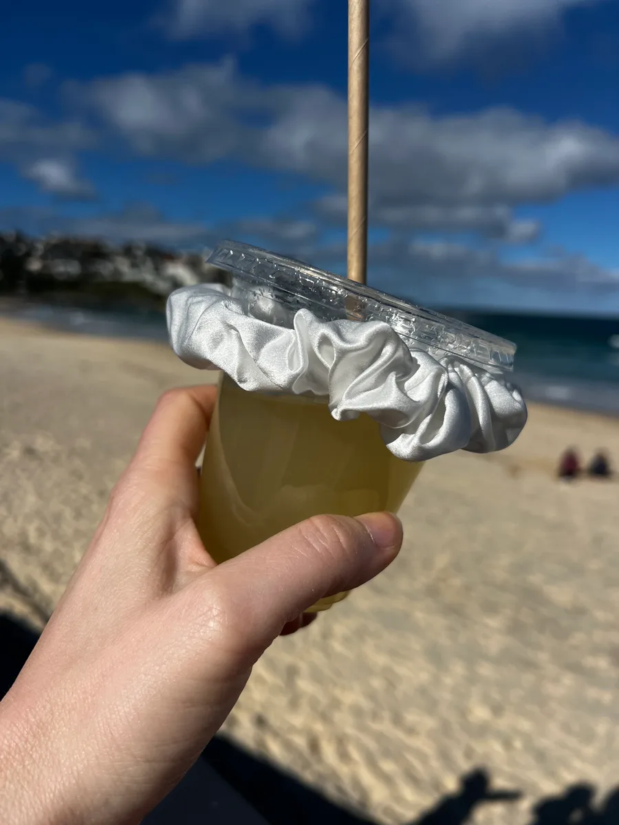 A hand holding a cold beach drink topped with a white Kova Silk scrunchie, ocean and sand behind