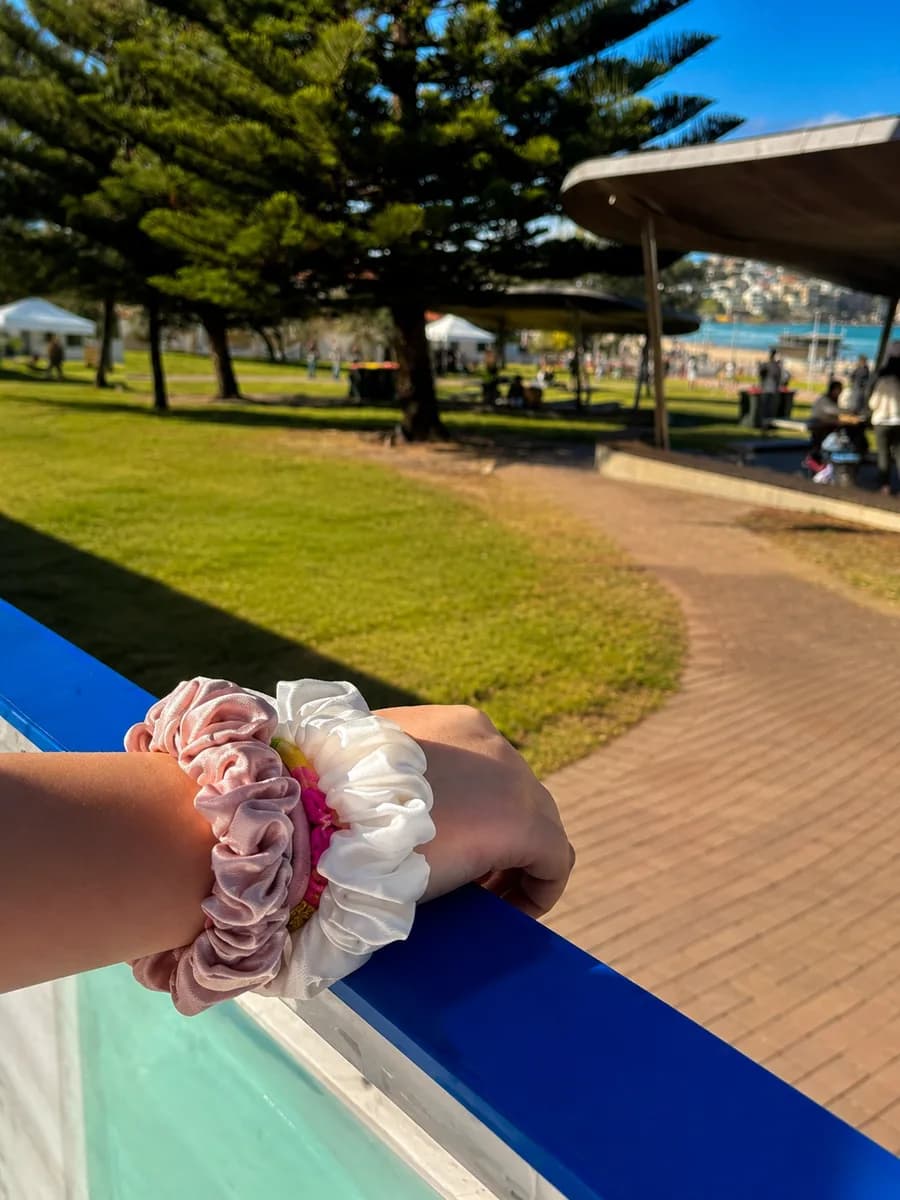 A wrist resting on a railing wearing pink and white Kova Silk scrunchies at a sunny beachside park