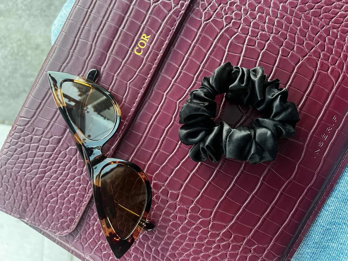 A black Kova Silk scrunchie and tortoiseshell cat-eye sunglasses arranged on a burgundy croc clutch
