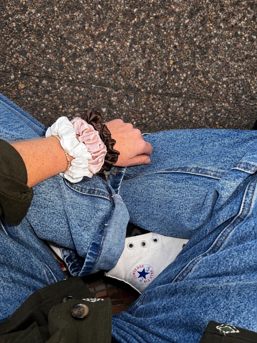 A wrist stacked with white, pink and brown Kova Silk scrunchies above white Converse sneakers